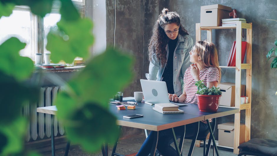 Two women collaborating at a desk in a stylish modern office with plants and a laptop.